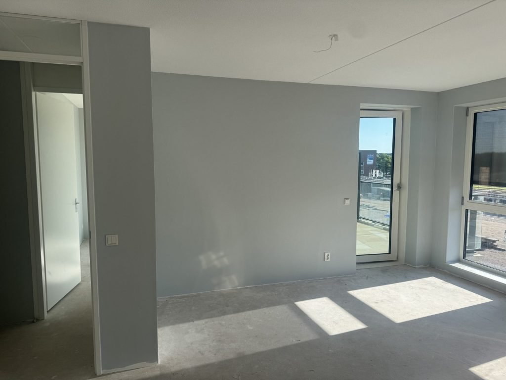Lege woonkamer met lichtblauw gesausde wanden en een balkondeur, de vloer nog kaal beton