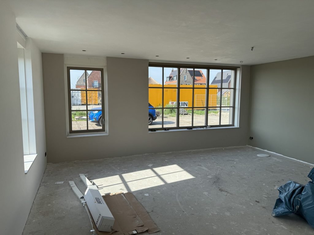 Woonkamer met grijsgroene wanden en een grote roedenpui die uitkijkt op een rij bakstenen huizen