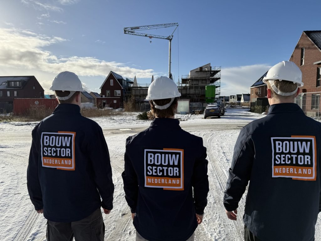 Drie vakmensen van Bouwsector Nederland met witte helm en logo op de rug bij huizen in aanbouw in de sneeuw